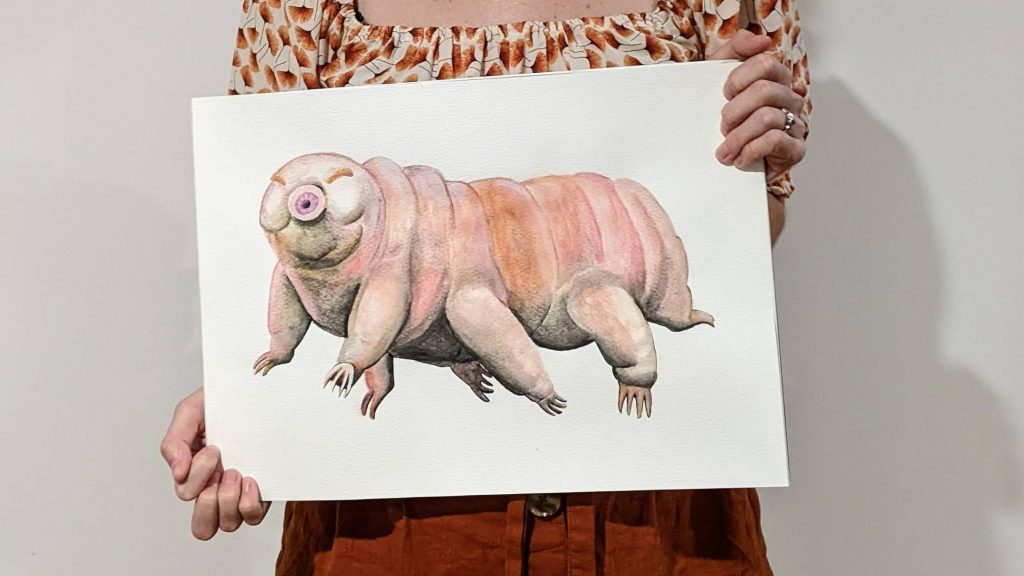 alise holding a painting of a watercolour tardigrade on an A3 sheet of paper.