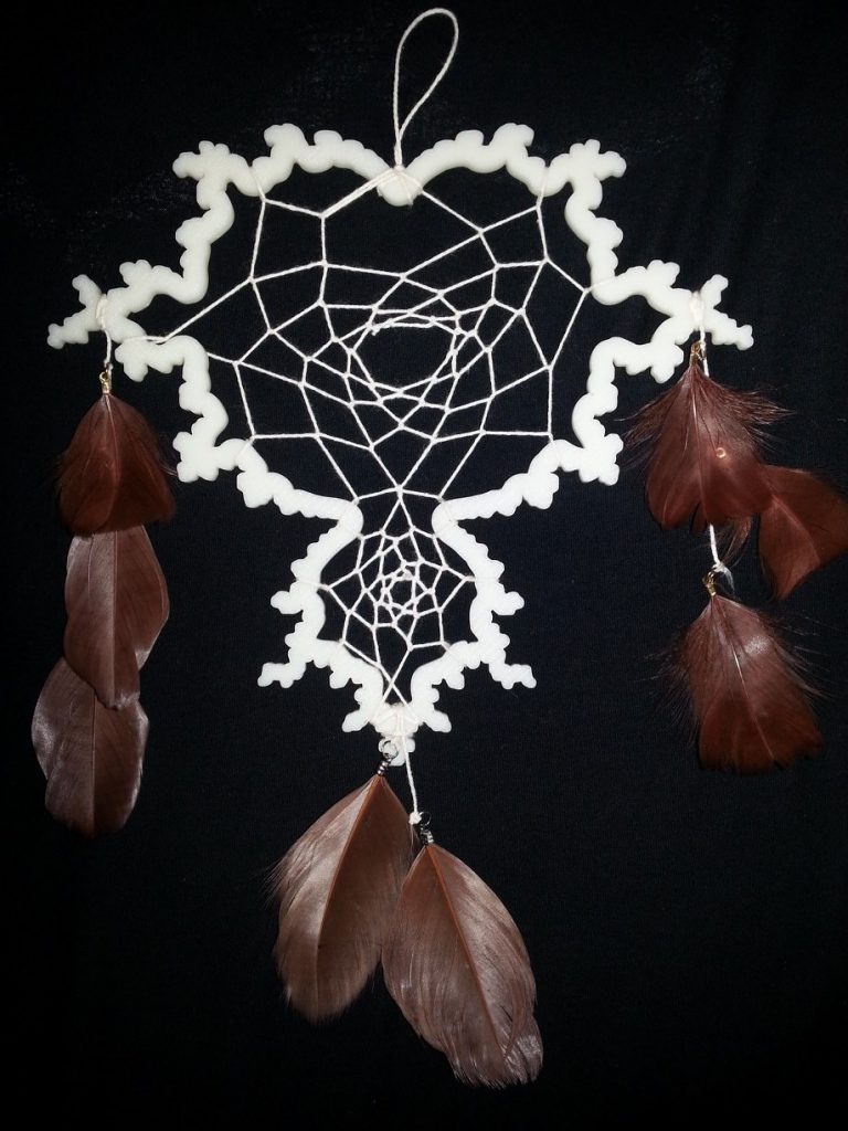 A white 3D printed outline of a Mandelbrot Set (fractal) on a black background. The shape has been decorated to look like a dream catcher, with strings and feathers. 